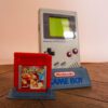 Photo d'une Game Boy Classic Grise sur son socle, vue de 3/4, avec le jeu Pokémon Rouge